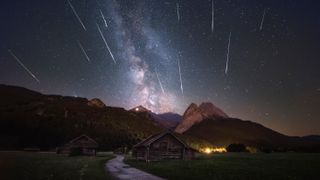 meteors streak through a star filled sky above mountains and small wooden lodges.