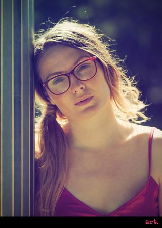 A portrait image of a woman with pink glasses and a pink dress, with her hair in a side ponytail standing next to a green pillar, taken with the Olympus Vintage I Art FIlter