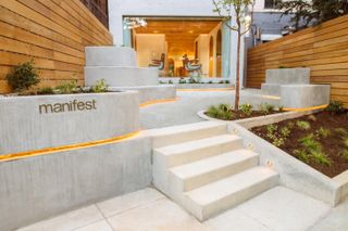 Concrete pavement with short staircase leading to entrance of shop and featuring landscaping with green plants