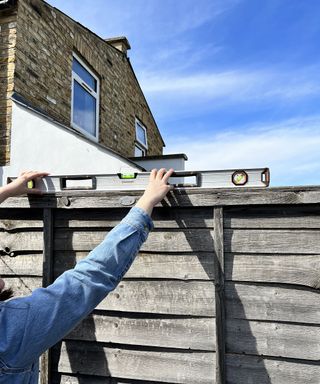 A female wearing blue denim jacket using spirit level guide on top of fence with house in background