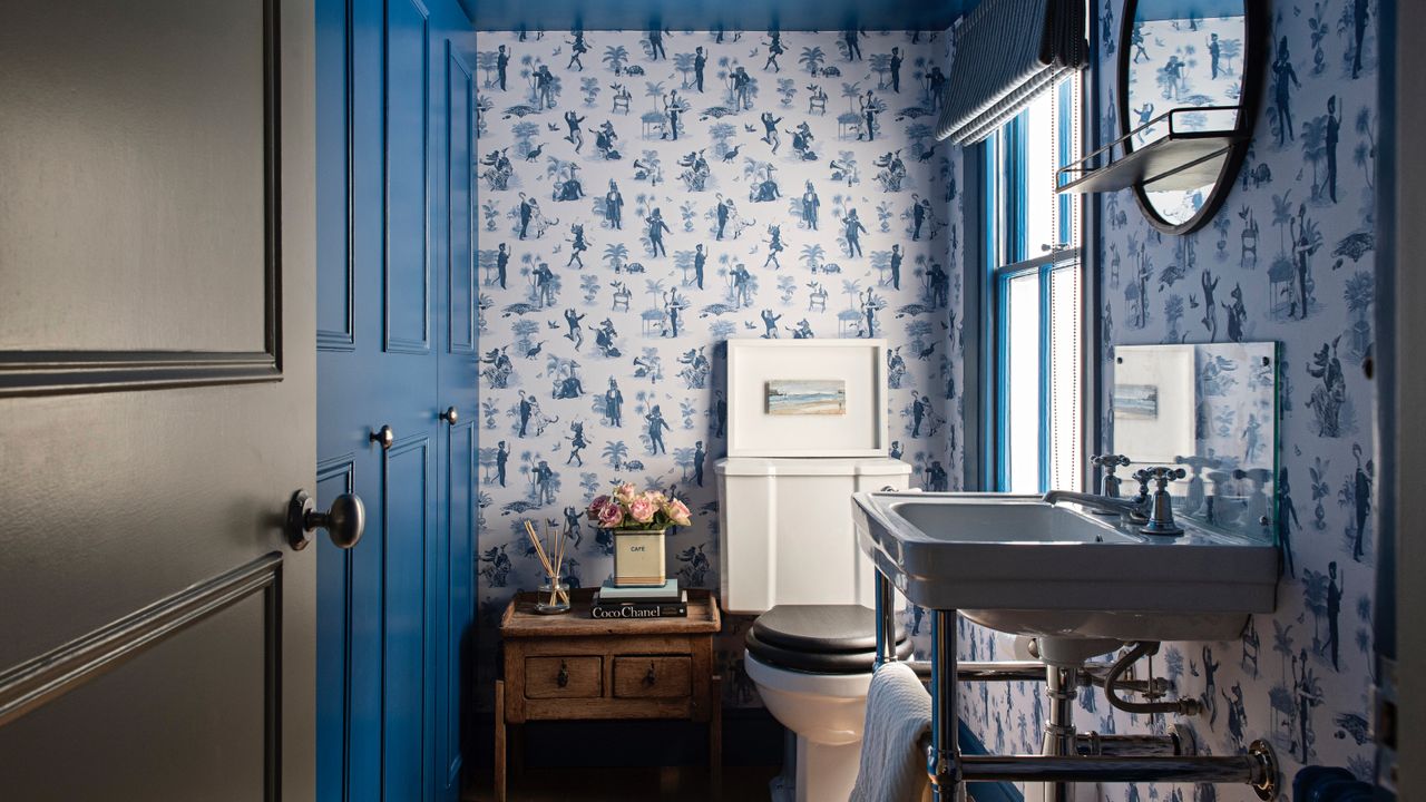 blue bathroom with blue wallpaper, mirrors, blue storage cabinet, cabinets on left, blue woodwork and ceiling, side table with flowers, basin with towel storage, blind