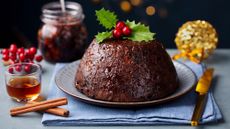 a traditional christmas dessert christmas pudding with a holly leaf and cinnamon sticks and mulled cider beside it, with a jar of mincemeat in the background