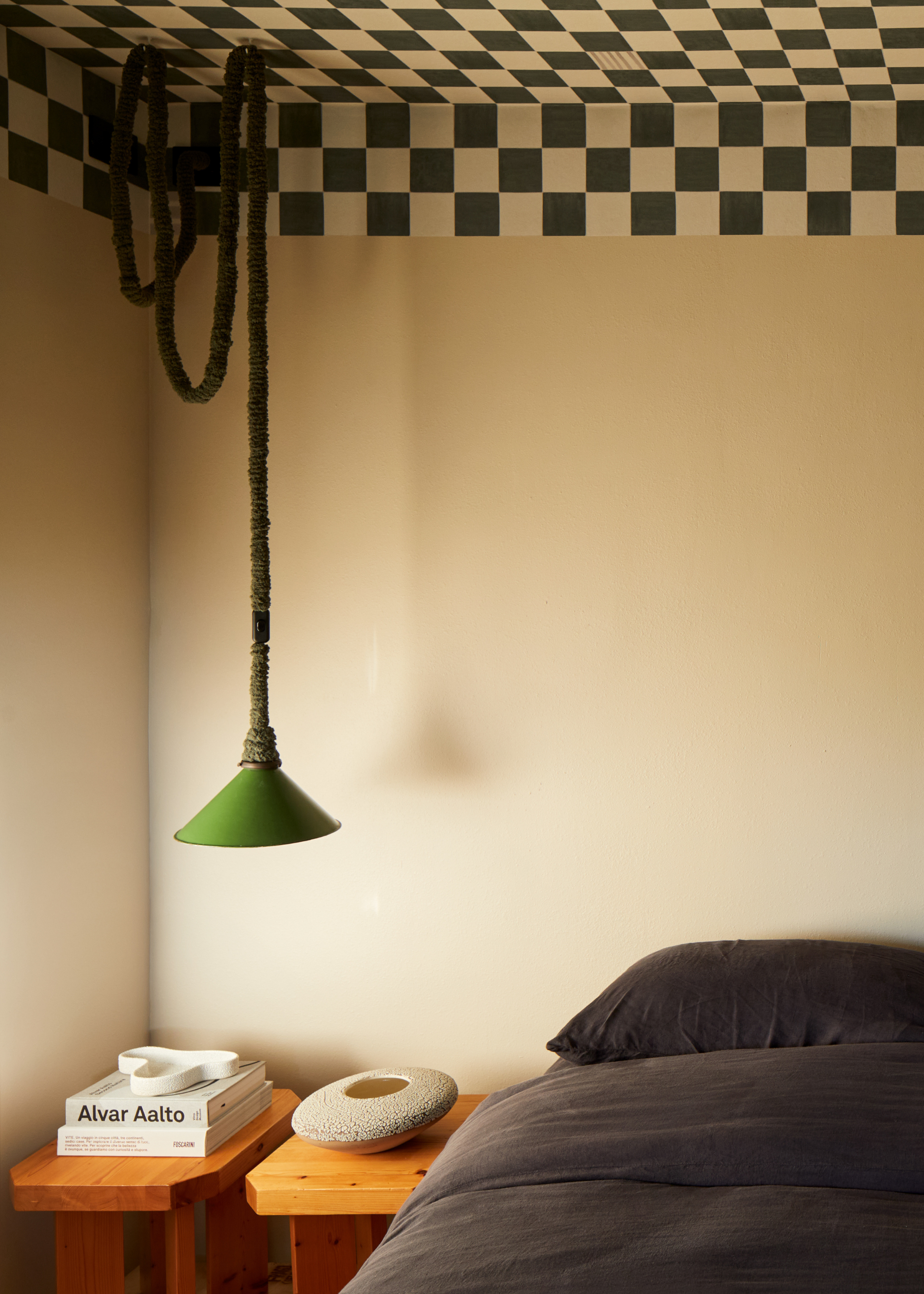 An off white bedroom with a dark bed, a pair of wood stool side tables, a looped ceiling pendant lamp by the bed and a deep green checkerboard ceiling