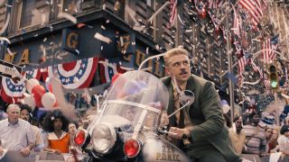 Still from the movie Indiana Jones and the Dial of Destiny. A man with short blond hair and wearing a suit is riding a New York police motorcycle, searching through a parade. There are American flags hanging everywhere and red, white and blue confetti and streamers about.