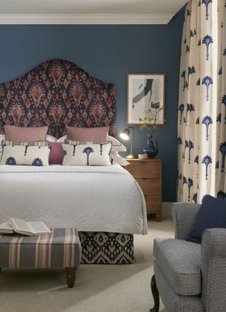 blue bedroom with blue print drapes and cushions, blue and red upholstered headboard, pink cushions, carpet, side table, footstool and blue armchair, artwork