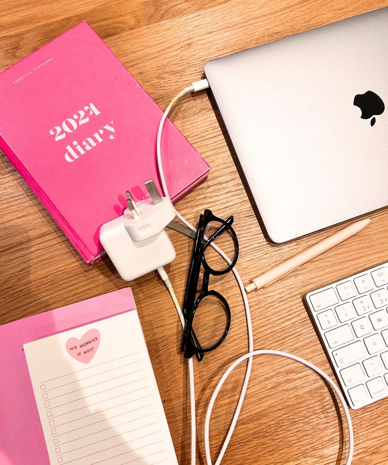 A messy wooden table before tidying work items away with a 2024 pink diary, notepad, wires, silver laptop and keyboard