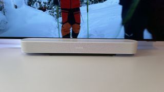 A white Sonos Beam Gen 2 soundbar photographed on a white desk in front of an OLED TV