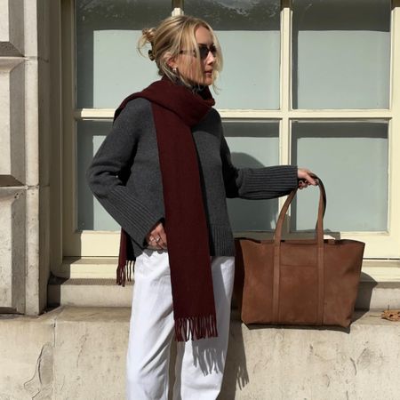 British style influencer Chloe Butler poses in London wearing a gray oversize sweater, burgundy scarf, brown suede tote bag, white jeans, and black loafers
