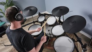 Over the shoulder view of a man in a black t-shirt playing an Alesis Strata Prime drum set in a blue-walled room