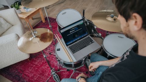 Man sits at a drum kit viewing Drumeo on his laptop