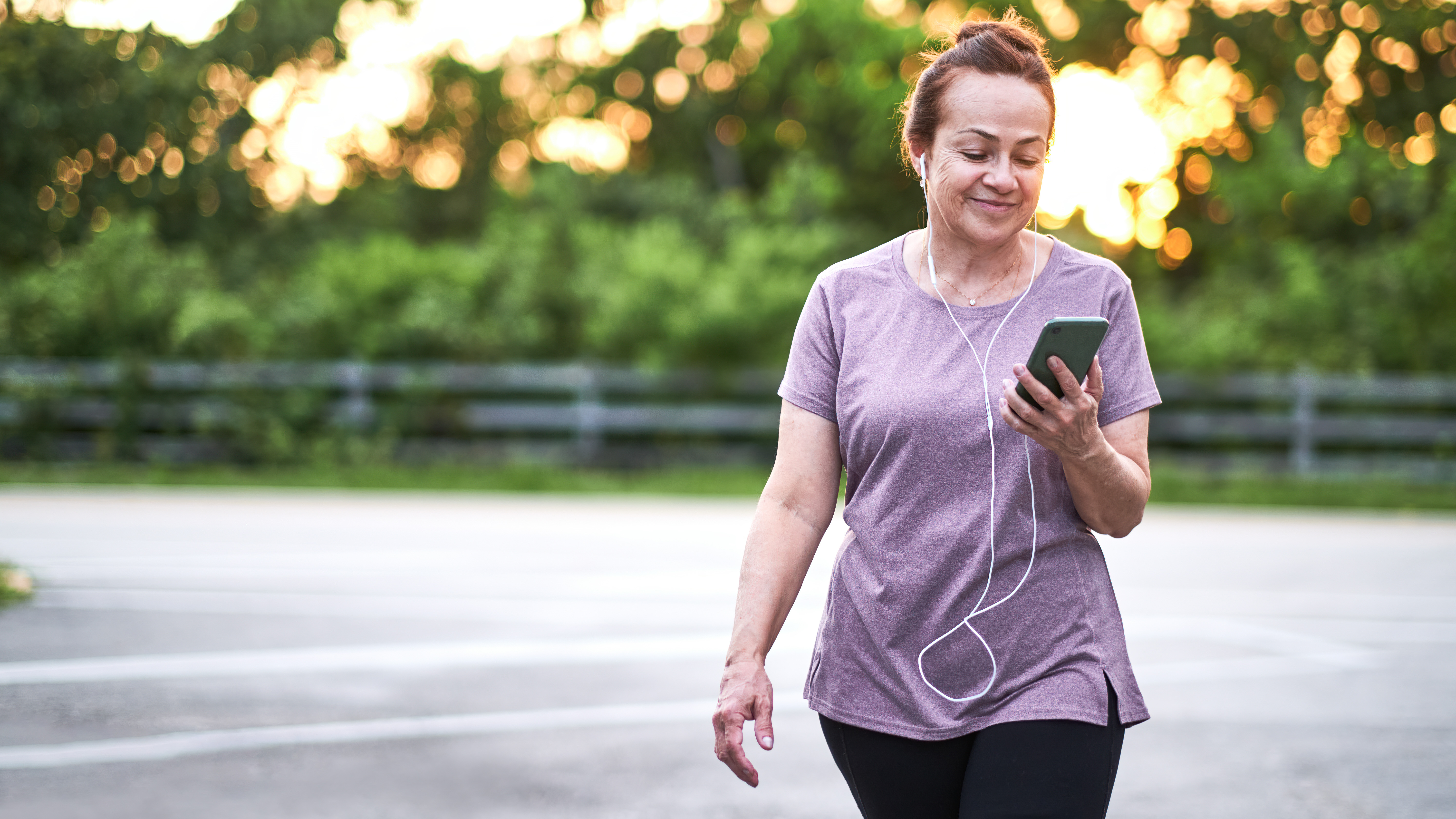 A woman walks along a paved surface looking at her phone with her headphones plugged into the device. She is smiling. Behind her we see a metal fence and bushes.
