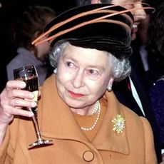 Queen Elizabeth wears an orange coat and raises a glass of champagne