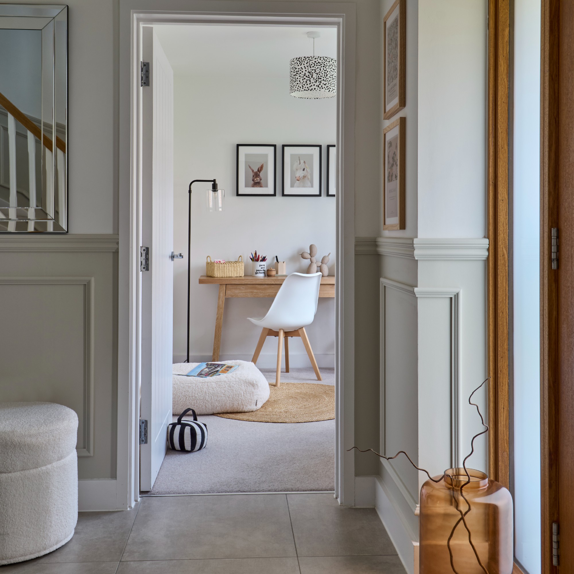 A neutral hallway with a dado rail and a white boucle storage stool with an open door into a home office