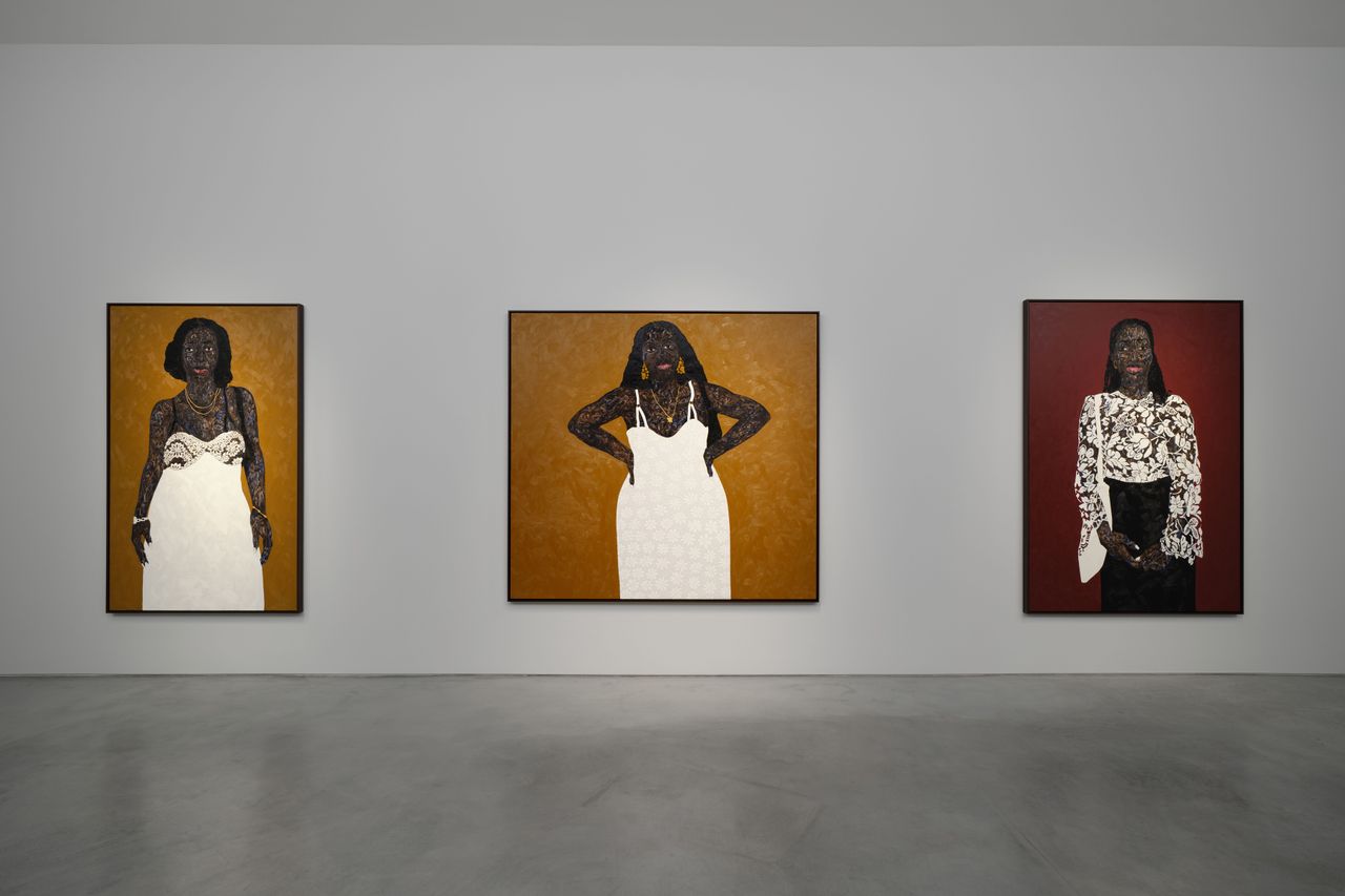 View of an art gallery installation with dark wood walls and multiple expressive portrait paintings. In the foreground, a large black metal structure with vertical bars frames additional paintings inside, while more portraits hang on the walls in the background.