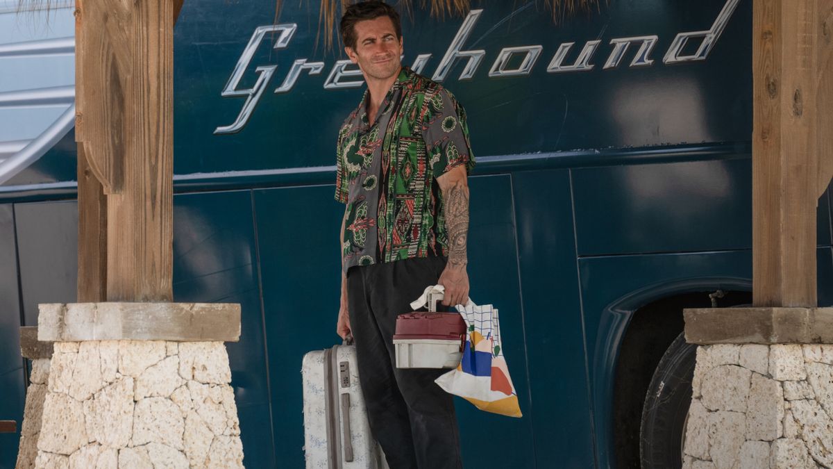 Jake Gyllenhaal stands smiling in front of a Greyhound bus in Road House.