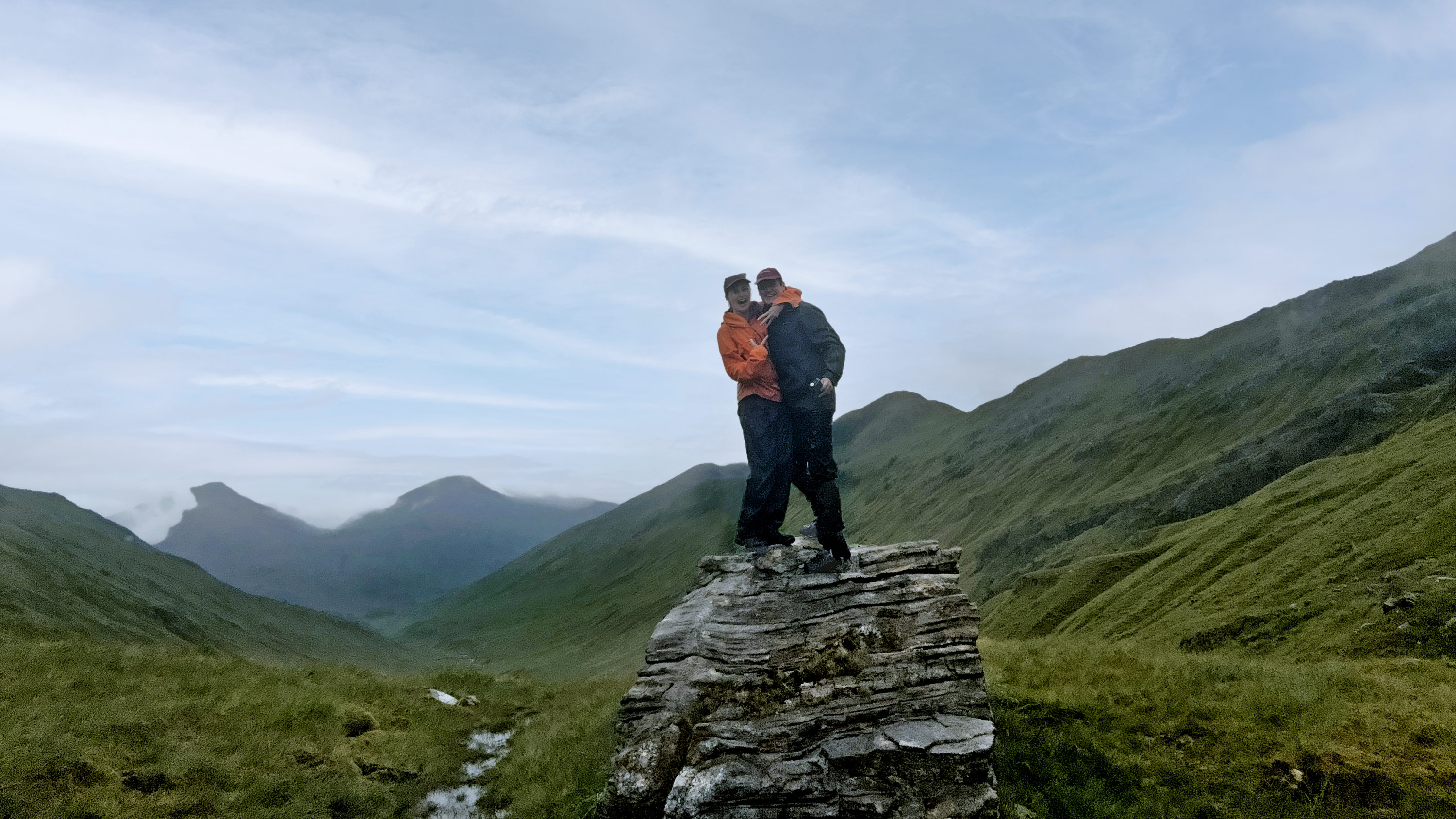 Two people stand on top of a pile of rocks, with mountain ranges in the background, the woman is pointing at her ring finger