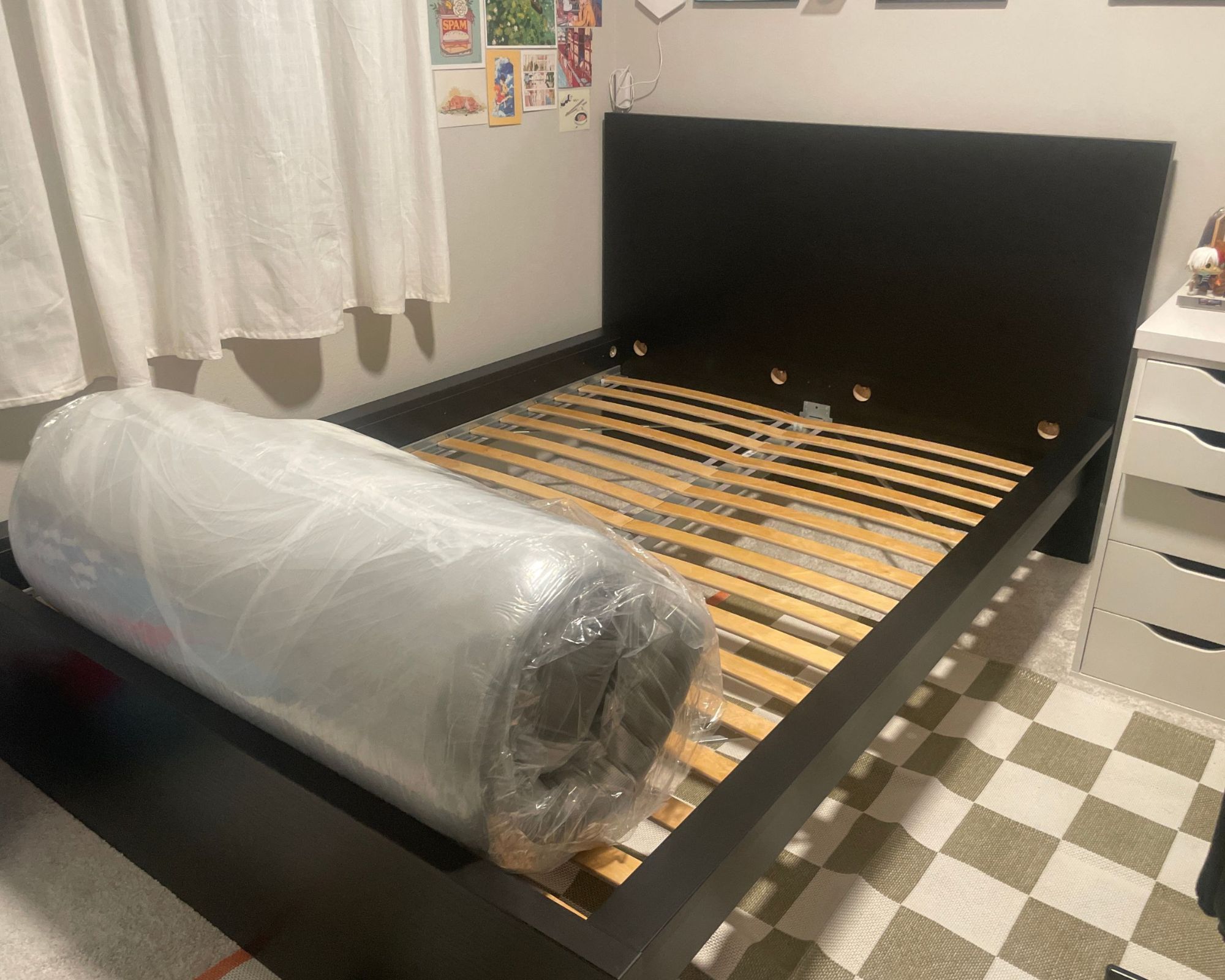 The WinkBeds WinkBed Mattress shown rolled on a black bed in a room with white walls, white curtains, and a white and grey checkered rug.