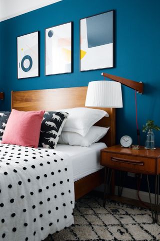 Bedroom with deep blue walls, deep mid century wooden bed frame and bedside table, white and black bobble bed covers and pink cushion