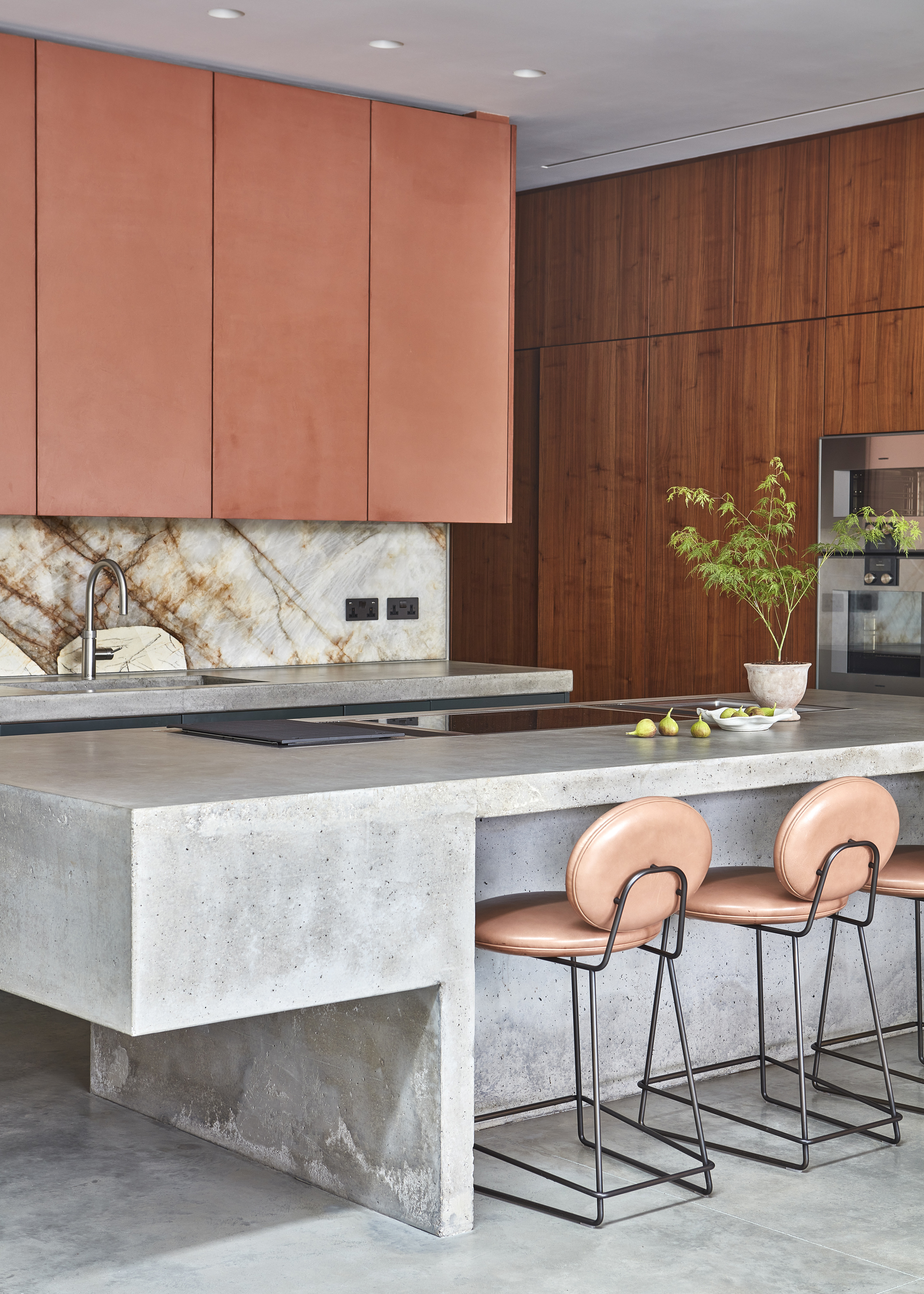 A modern kitchen with a concrete kitchen island, pink bar stools, and pink upper cabinetry