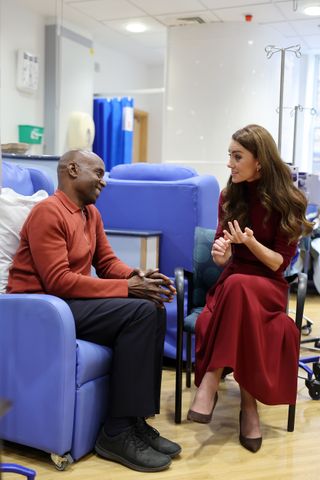 Kate Middleton wearing a red skirt and sweater sitting in a chair talking to a man wearing an orange top sitting in a chair at Royal Marsden hospital