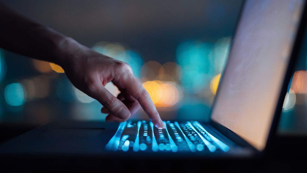 A man uses his index finger to type on a laptop, only his hand showing.