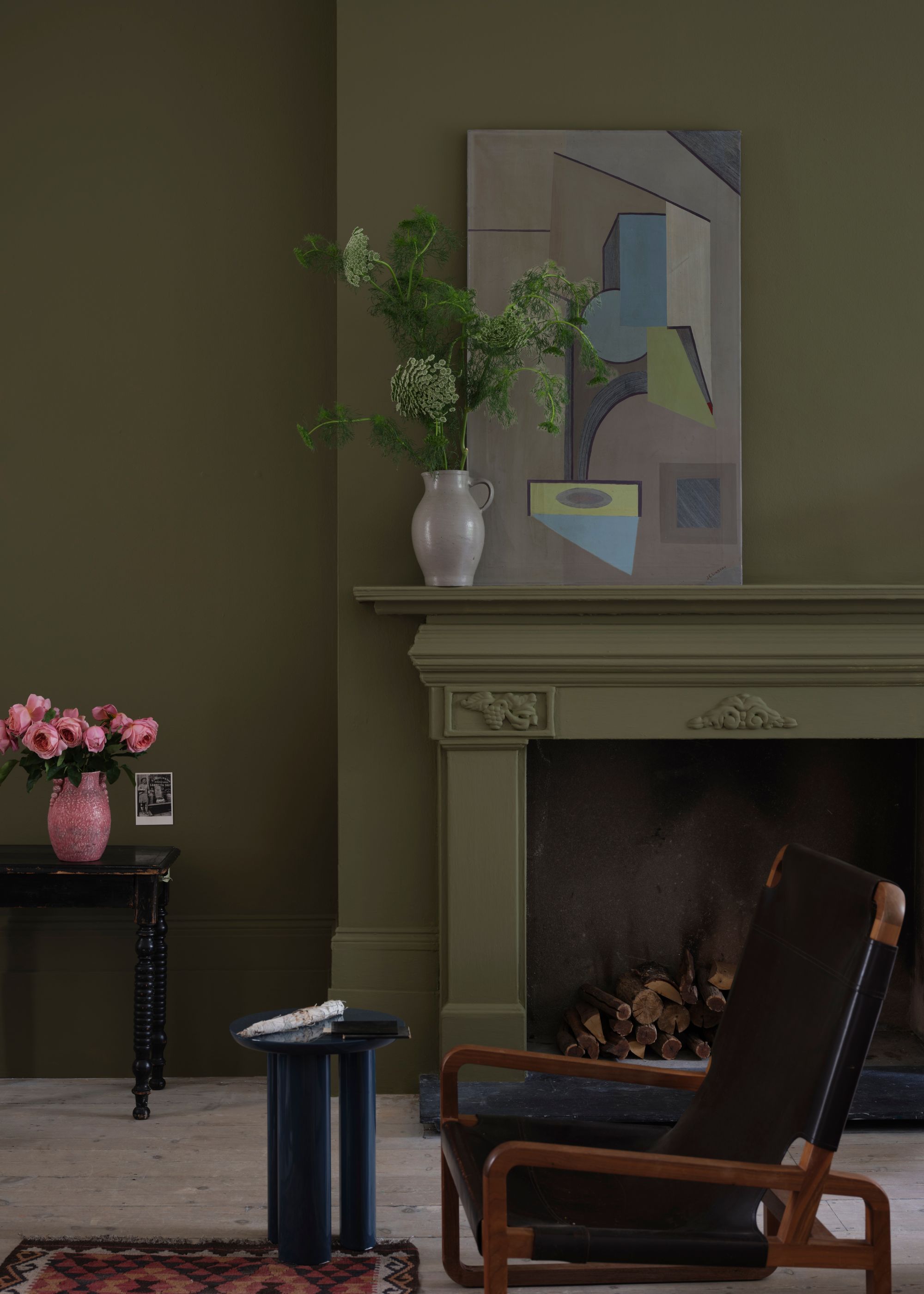 A cozy living room with dark olive green color-drenched walls and fireplace, a black armchair, a black side table with a vase of pink flowers, and a geometric artwork displayed on the fireplace.