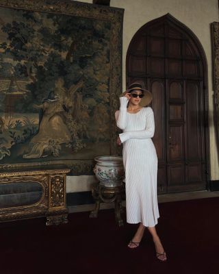 woman wearing white rib-knit long-sleeve midi dress with oversize straw sun hat, black sunglasses, and black strappy sandals in front of a wall tapestry