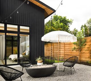 outdoor seating area in garden with gravel courtyard, timber fence, umbrella, fire pit and house with black exterior