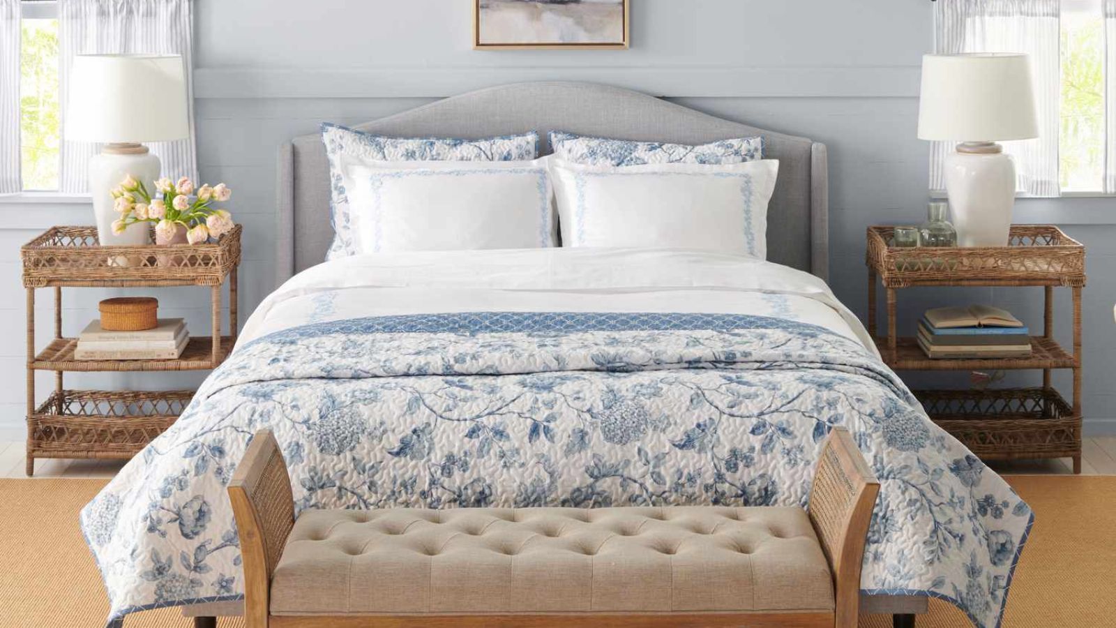White and blue floral sheets on a bed against blue walls behind a chaise lounge, with a wooden nightstand either side. 