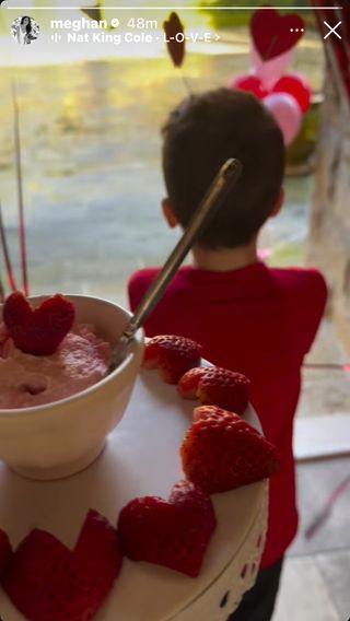 Prince Archie wearing a red shirt facing a back door with a tray of pink heart shaped strawberries in front of him