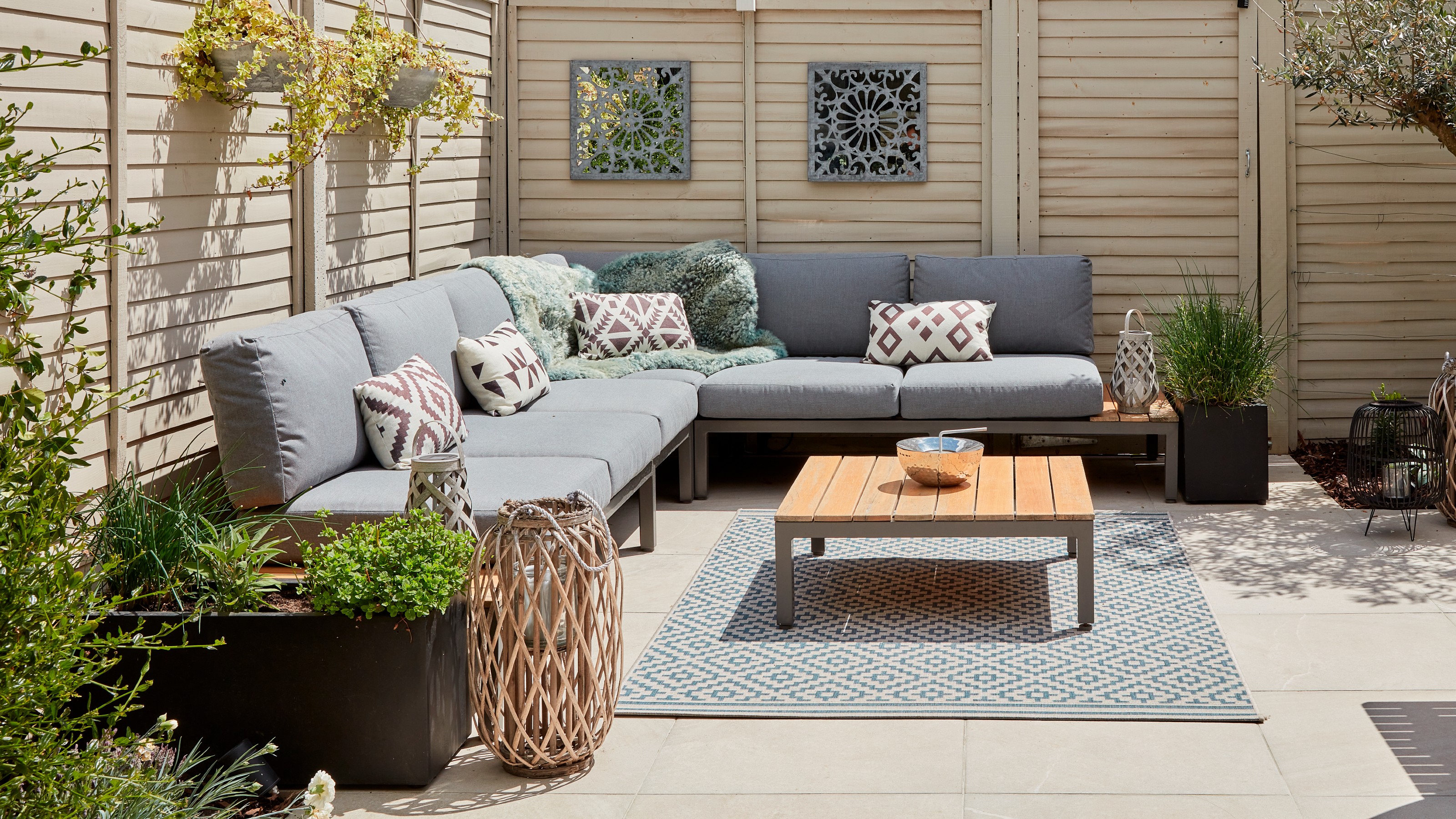 A patio with a grey corner outdoor sofa and a square coffee table in the centre on top of a patterned rug