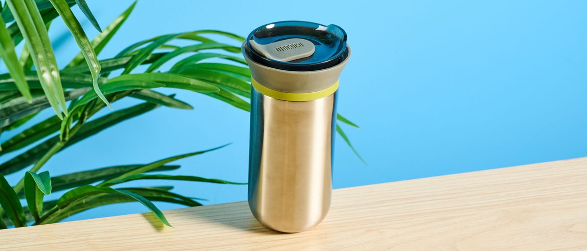 Wacaco Cuppamoka, a stainless steel coffee pour-over brewer, photographed against a blue background, with brown filters and a sippy lid