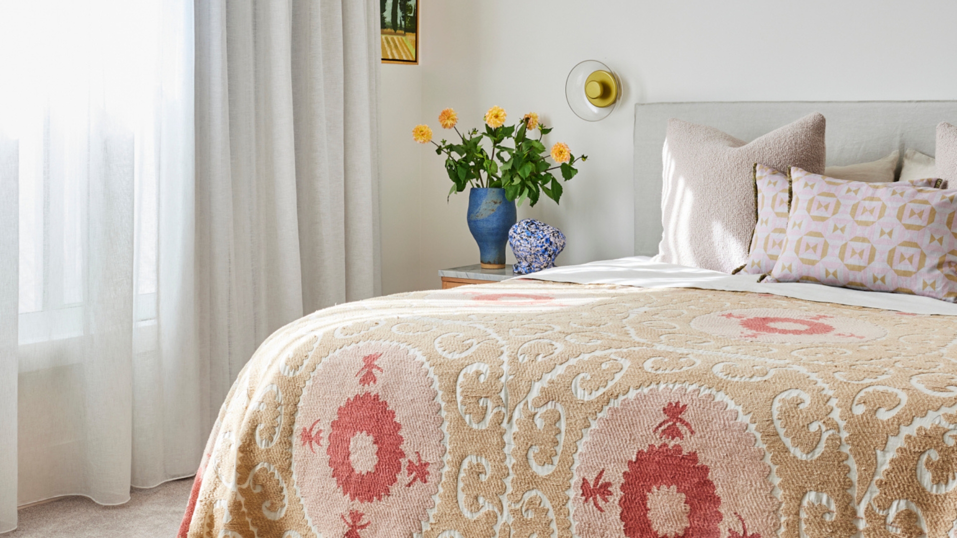 A white bedroom with a patterned quilt, patterned pillows, a blue vase of flowers, and a painting