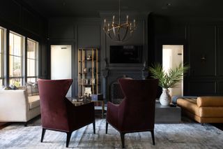 A living room with black walls and ceiling, two deep burgundy wingback chairs, and large windows