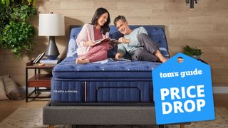 A man and a woman laugh while reading a magazine on top of the Stearns & Foster Lux Estate Mattress with a blue price drop image overlaid
