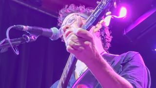 Jason Newsted playing an acoustic guitar onstage in 2026