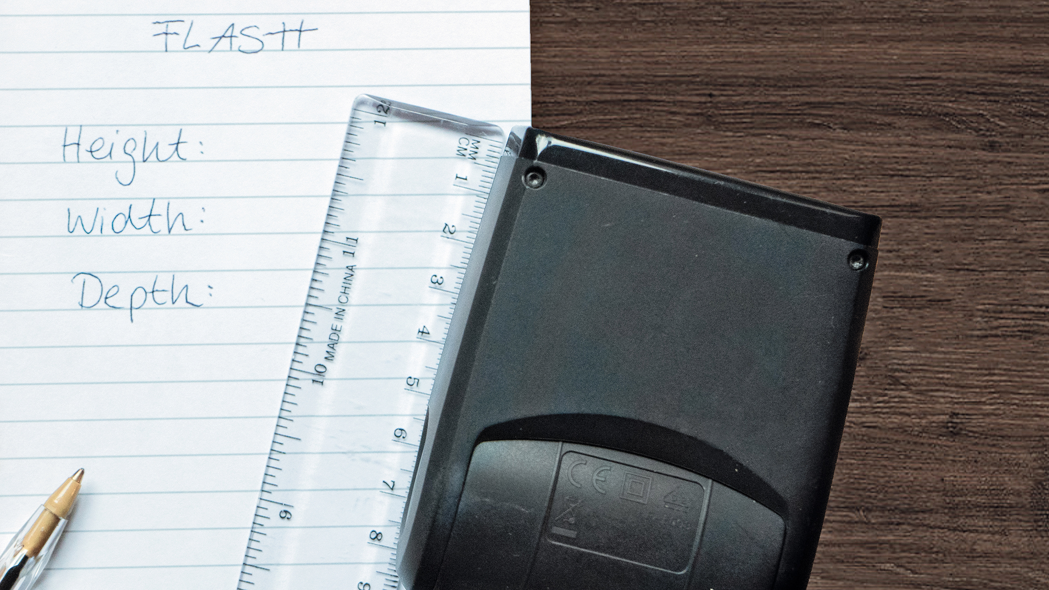 A black flash device rests on lined paper with measurements labeled: Height, Width, and Depth, alongside a clear ruler and a pen