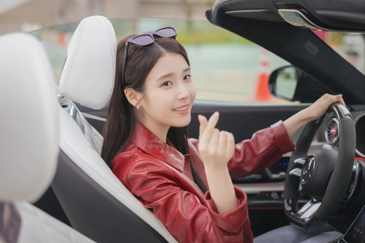 IU as Seong Hui-ju, making a Korean finger heart as she sits in the front seat of a convertible, in 'Perfect Crown'