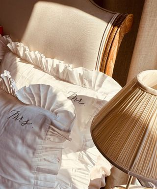 A birds eye close up view of a beige bed with white ruffled pillows with 'mr' and 'mrs' stitched onto the corners in black writing and a beige lampshade with a brushed gold base next to it