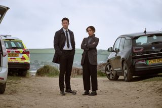 DI Matthew Venn (Ben Aldridge) and DS Jen Rafferty (Pearl Mackie) stand on the beach looking into the camera lens, surrounded by police cars