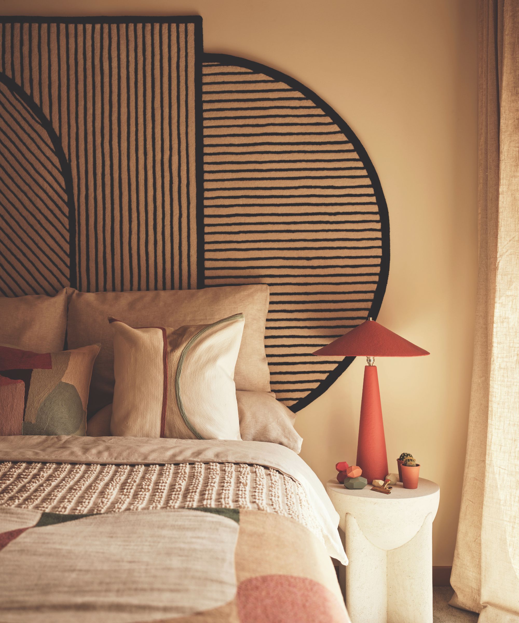 A contemporary bedroom designed by Tola Ojuolape Studio featuring a bold, custom upholstered headboard with a black-and-white striped semicircular motif. A sculptural red table lamp sits on a stone side table next to neutral linen bedding and a textured geometric throw.