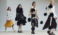 Four women on a catwalk, one in a shirt and yellow skirt, one with a back coat with fur cuffs and hem, and two in a shirt and black skirt with a scarf featuring large black fur pompoms