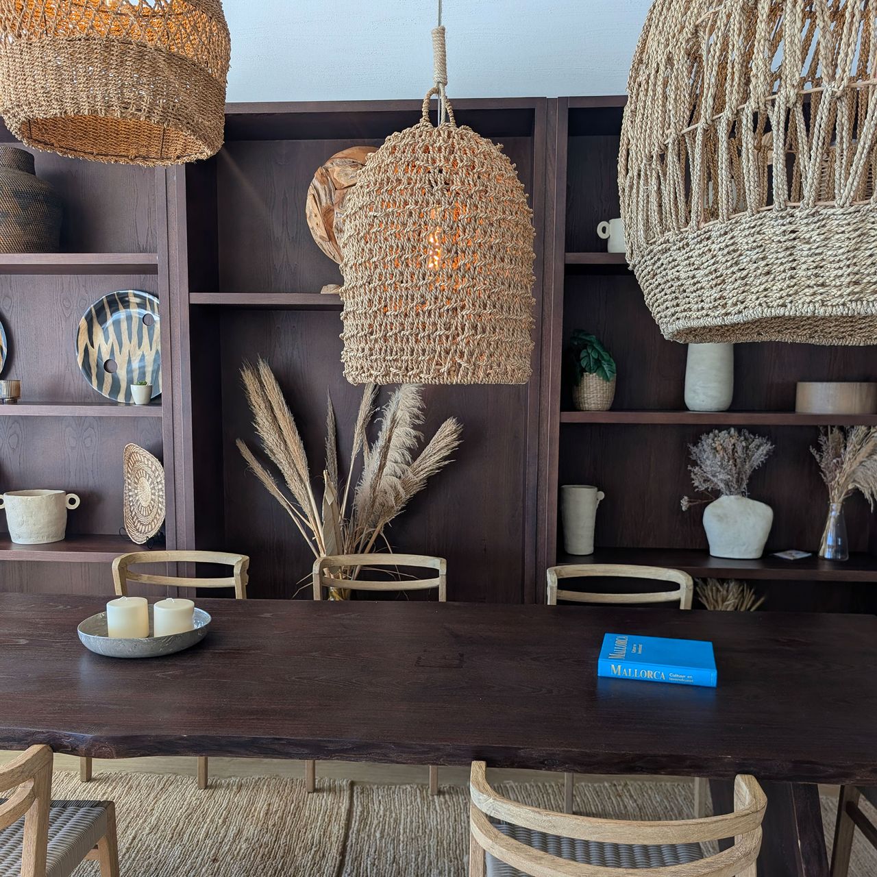 Dark wood dining table topped by woven lampshades in front of built-in dark wood floor-to-ceiling shelving