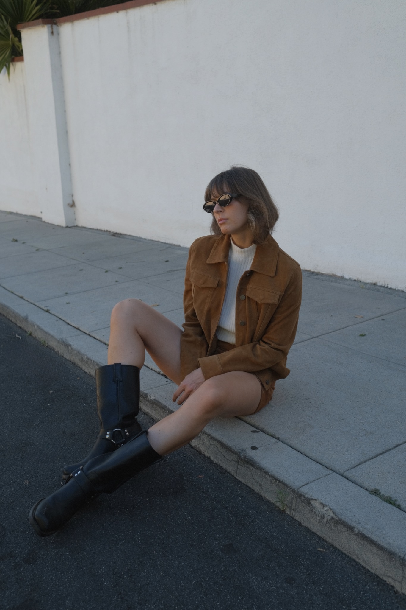 Kathryn Zahorak wearing sunglasses, white turtleneck, suede jacket, suede shorts, black moto boots