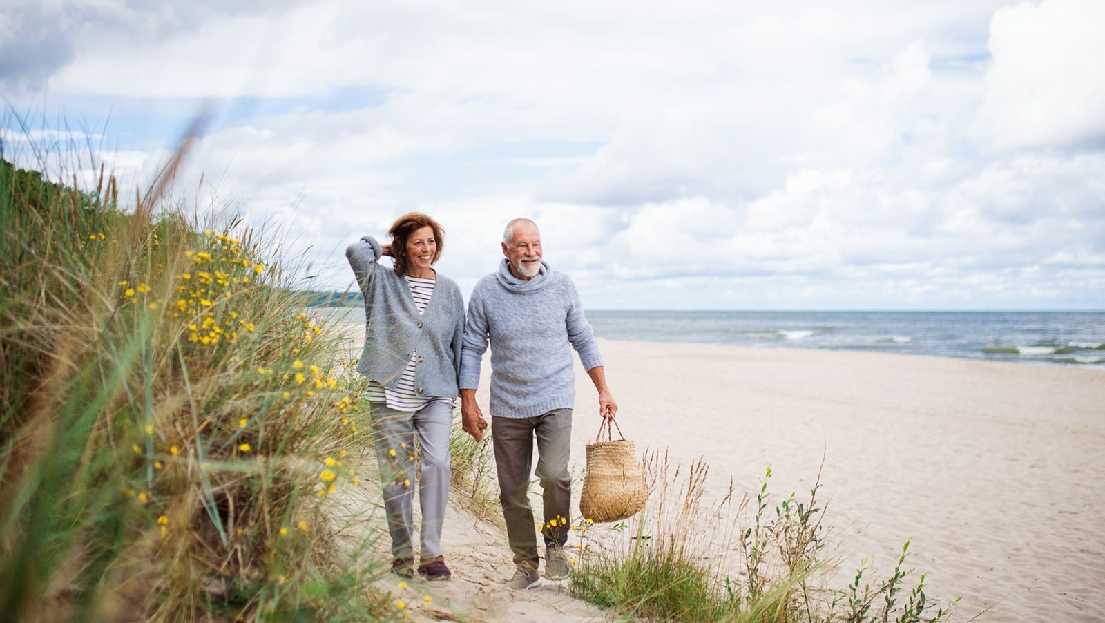 Happy senior woman and man holding hands and walking outdoors on sandy beach in autumn.