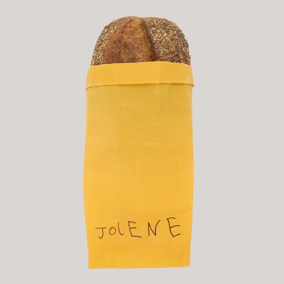 A resistant paper bread bag in yellow with the handwritten logo "JOLENE" holds a loaf against a pale grey background.