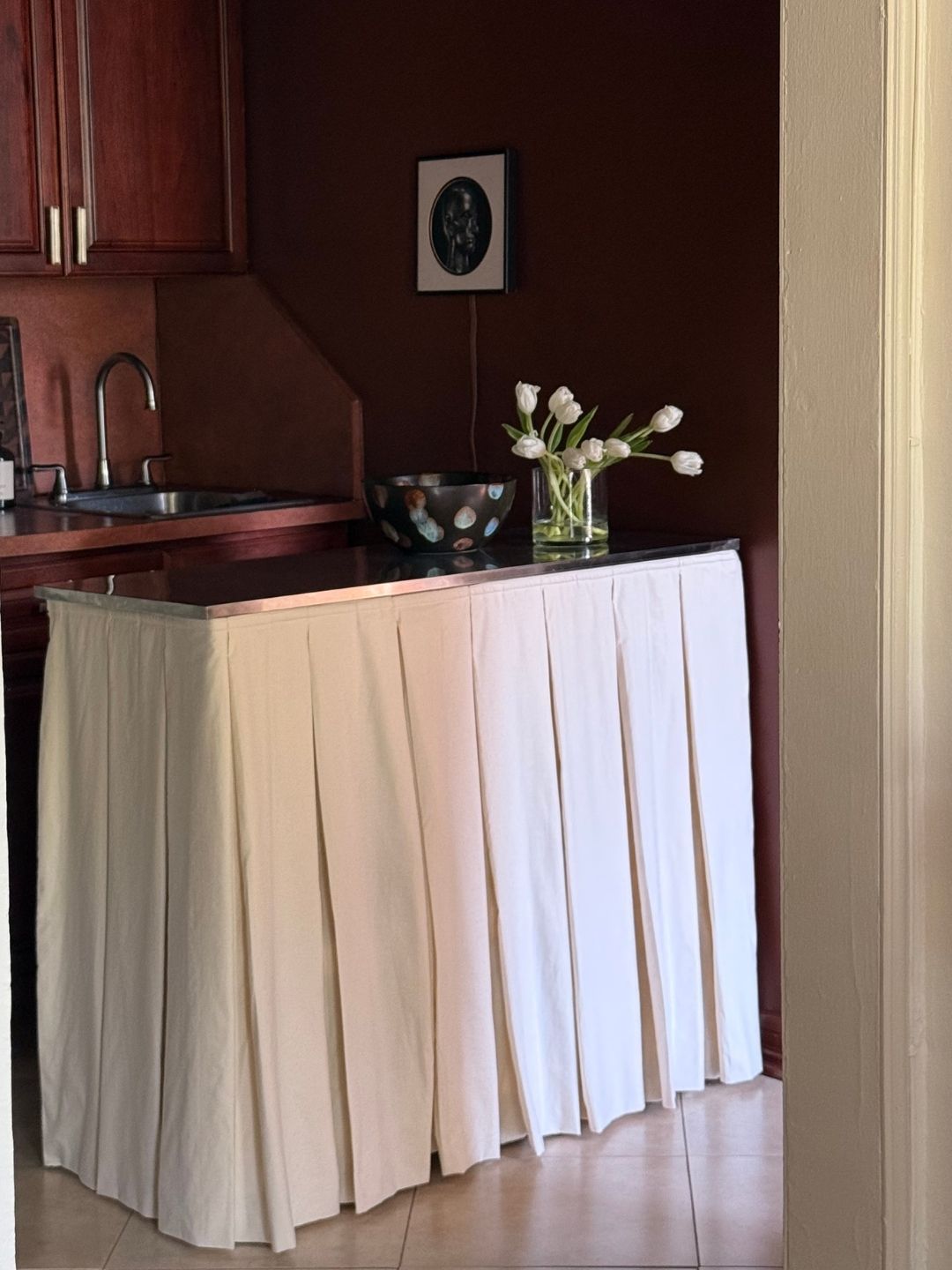 Image of a dark reddish-brown kitchen with a small kitchen island that has a chrome top and a white box-pleat skirt.
