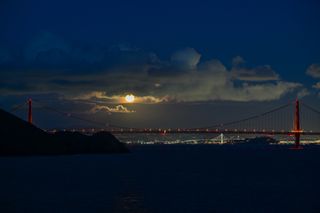 In the center of the image the full moon shines through the clouds and the partly illuminated Golden Gate Bridge sits below.
