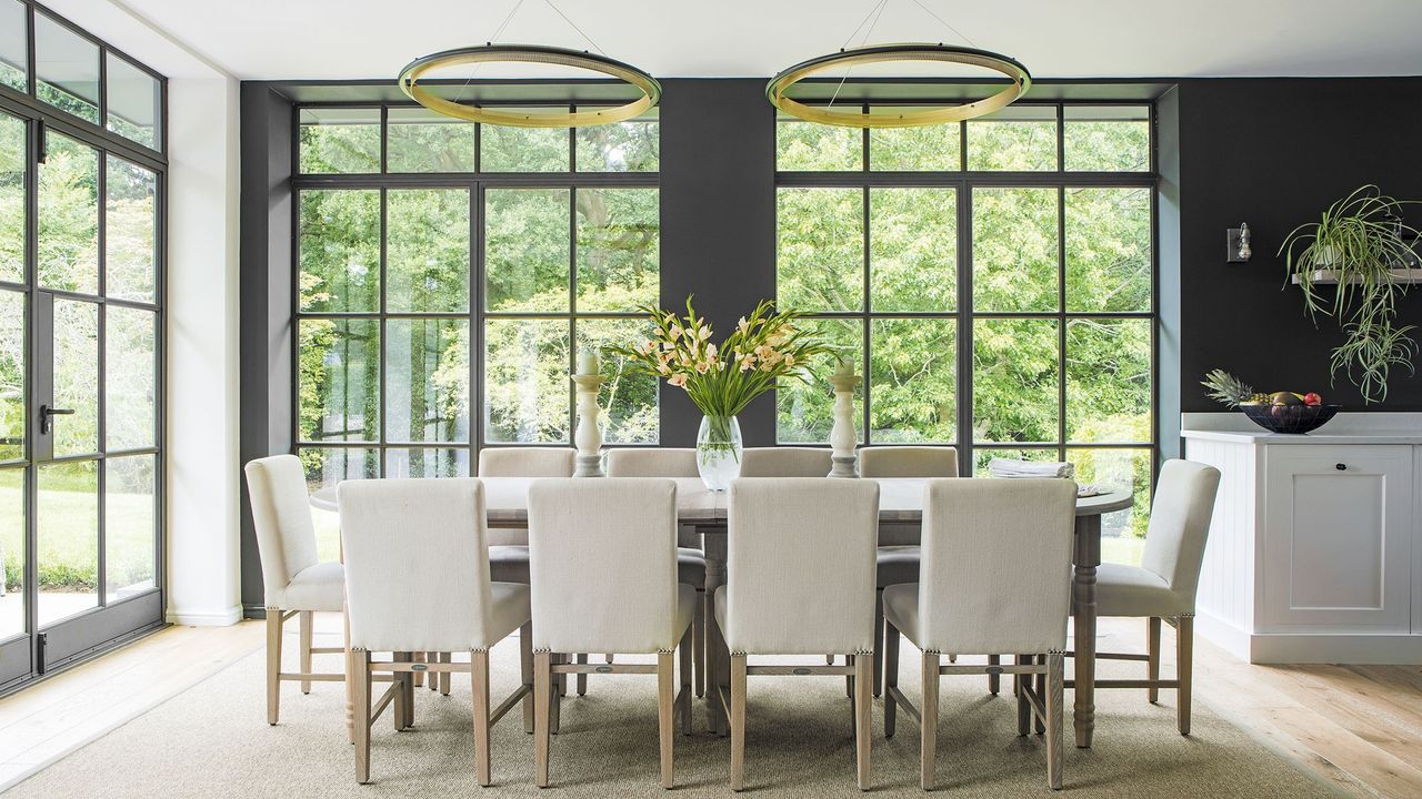 large dining are with floor to ceiling panel windows with black frame, a large 10 seater dining table in the middle with white chairs and two statement ceiling drop ring light fixtures above