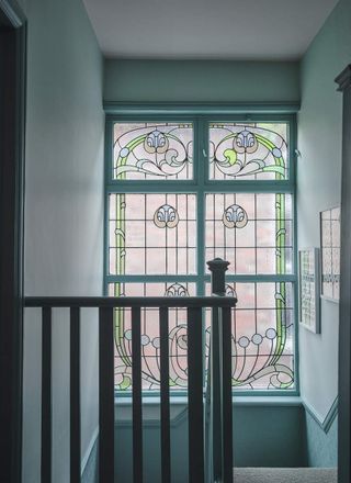 Image of a teal staircase with a large stained glass window. The design is Art Nouveau-inspired and the color palette is green and light purple.